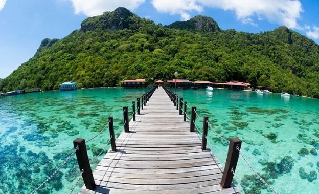 A stunning wooden jetty stretches over crystal-clear turquoise waters, leading towards lush green hills. A perfect tropical escape showcasing Malaysia’s serene island beauty.