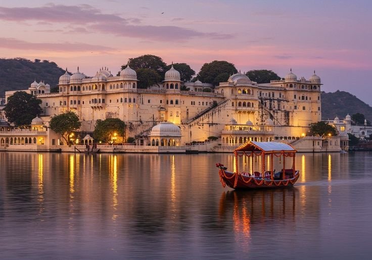 Illuminated Udaipur City Palace reflected in Lake Pichola at sunset with a decorated boat in the foreground.